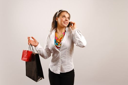 Modern looking young woman wearing white jacket talking on the phone and holding shopping bags