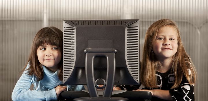 Two young girls looking from behind computer monitor