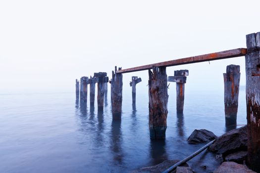 Photograph of a broken worn down boat dock, in the morning mist. Shore is cluttered with trash.