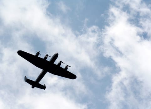 The silhouette of a Lancaster bomber against blue sky with white puffy clouds.