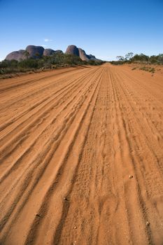 Dirt road with tire tracks in Uluru Kata Tjuta National Park, Australia with Mount Olga in the distance.