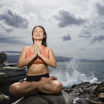 Asian woman sitting on rock by ocean in lotus pose with eyes closed in Maui, Hawaii