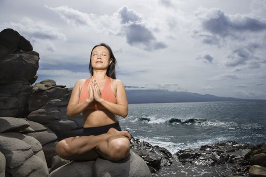 Asian woman sitting on rock by ocean in lotus pose with eyes closed in Maui, Hawaii