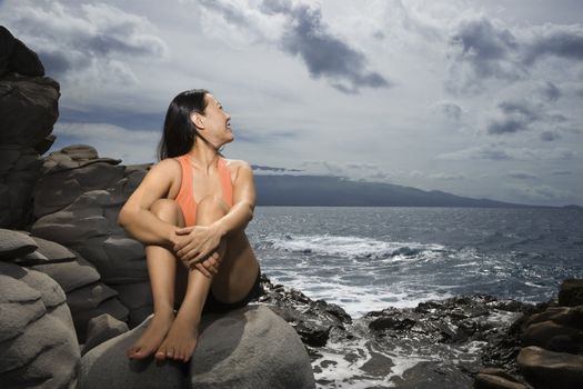 Asian woman sitting on rock by ocean looking over shoulder smiling in Maui, Hawaii