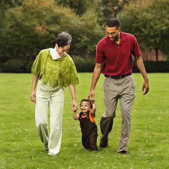 Mother and father helping toddler walk holding his hands in park.