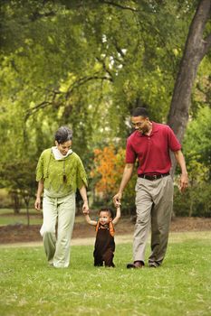 Mother and father helping toddler walk holding his hands in park.