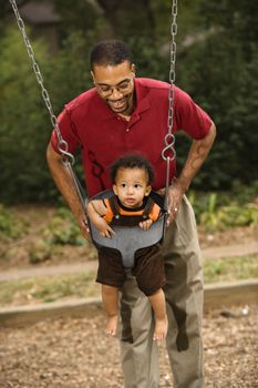Father pushing young son on swing in park in playground.