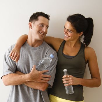 Man and woman at gym in fitness attire holding water bottles standing against wall smiling at eachother.