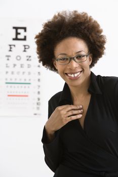 Portrait of woman with afro wearing eyeglasses with medical eyechart in background.