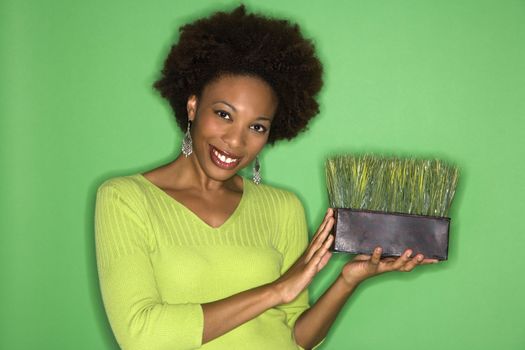 Smiling woman with afro holding pot of grass against green background.