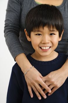 Portrait of Asian boy smiling with mother standing behind with arms around him.