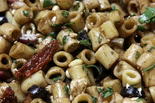 A close-up of greek pasta salad, featuring sun-dried tomatoes, black olives, red onion, pine nuts, spinach,feta cheese, balsamic vinegar and ditali pasta.
