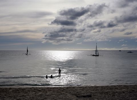 Beach on a sunset with silhouettes of sailing boat and bathing children