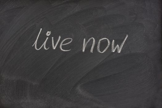 live now phrase handwritten with white chalk on blackboard with strong patterns of eraser smudges