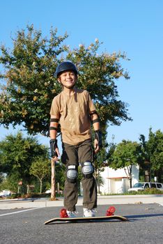 Teenage boy smiling with a skateboard on the ground on a sunny day with blue sky and trees in the background.