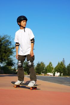 Teenage boy riding a skateboard on the sidewalk of a parking lot on a sunny day with blue sky and trees in the background.