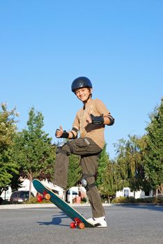 Smiling teenage boy with his thumbs up standing on a skateboard on a sunny day with blue sky and trees in the background.