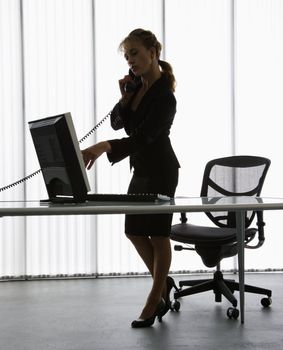 Silhouette of Caucasian businesswoman standing at computer desk on telephone.