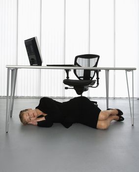 Caucasian businesswoman sleeping on floor under computer desk.
