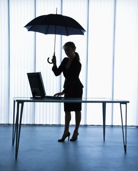 Silhouette of businesswoman standing at computer desk holding umbrella.