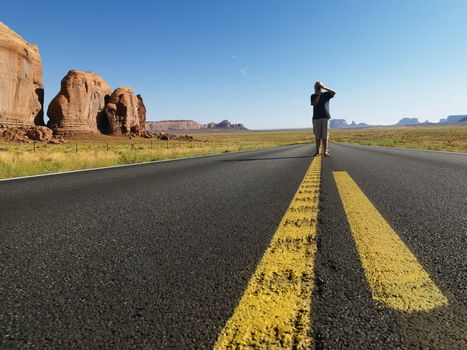 Caucasian teenage boy standing in open road in desert with landforms taking photograph.