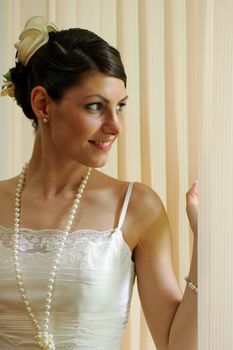 Smiling young bride in traditional white wedding dress peeping through blinds.