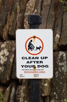 an old, worn, rust stained dog fouling sign on a black post in front of a stone wall, with fixed and maximum penalty removed.