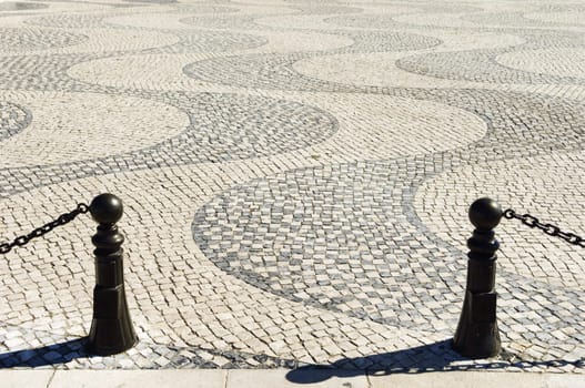 A view of a wavy cobblestone plaza with black security chains and poles in the foreground, Lisbon, Portugal