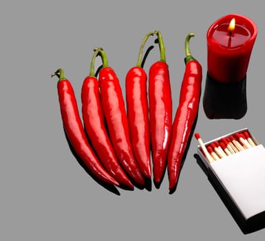 fresh red chili peppers  with matches and lighted red candle over grey reflective surface