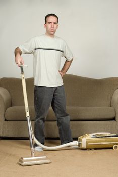 A full body view of a young man vacuuming the carpet and looking grouchy