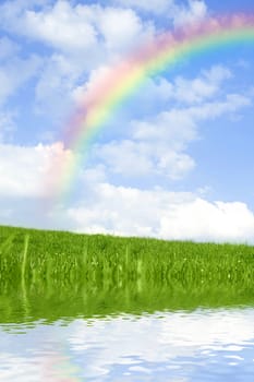 Beautiful green grass against blue sky and rainbow, with water reflection.