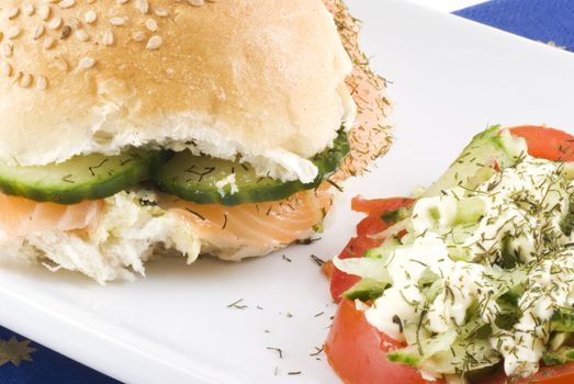 Close up of a sesame bun with salmon and cucumber with a tomato salad on the side.