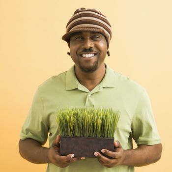 African-American mid-adult man wearing hat holding potted grass smiling at viewer.