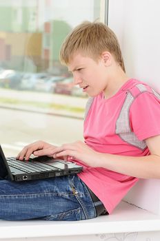 The joyful teenager with a computer sits on a window