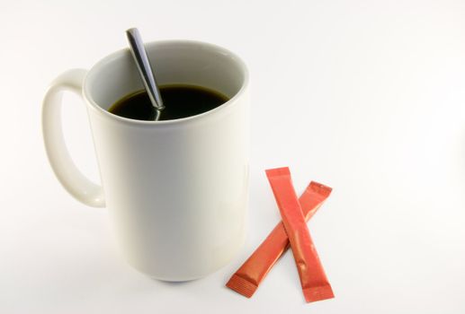 Tall white coffee mug with two red sugar sachets and spoon on a white background