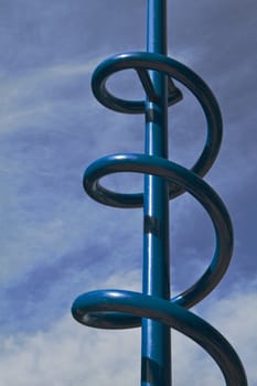 Sun lit blue metal climbing spiral feature of a childrens playground with cloudscape background