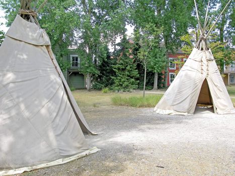Two teepees in a forest with buildings behind.