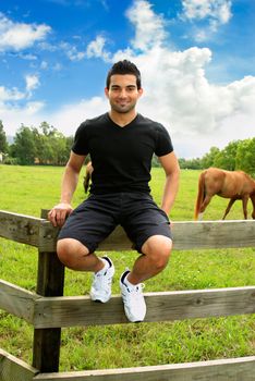 A smiling man sitting on a fence in rural countryside farm.