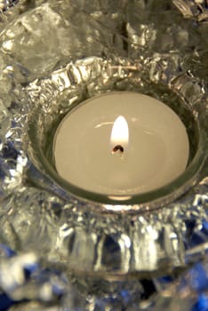 A single, white burning tea light candle with a  decorative, shiny background. Selective focus, vertical view.