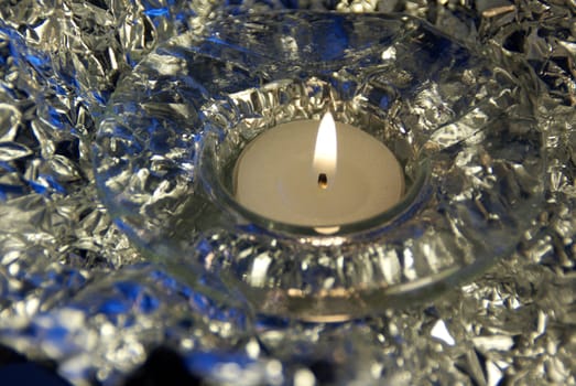 A single burning tea light candle with a decorative silver and blue background. Selective focus, horizontal view.