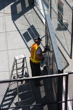 Worker Washing Window v1 shows a man wearing an orange vest with reflective stripes cleaning glass while listening to music from a portable stereo plugged in with an electical power cord plugged in nearby. 
