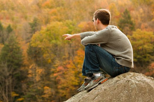 Young male hiker in jeans pointing while sitting on a boulder in front of fall leaves