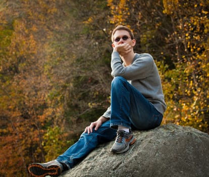 Young male hiker in jeans pondering while sitting on a boulder in front of fall leaves