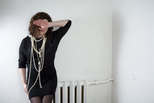 Young upset woman in pearls beads and black dress sitting on radiator closing eyes by hands. Woman`s problem