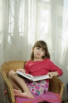 little cute serious girl seven years old reading book sitting in chair