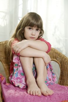 Little cute dreaming girl sitting on chair at window hugging her knees