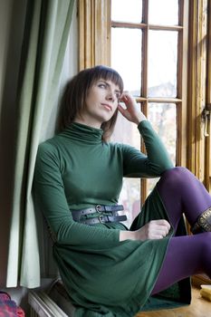 Attractive pensive woman in green dress and violet stocking sitting on window-sill