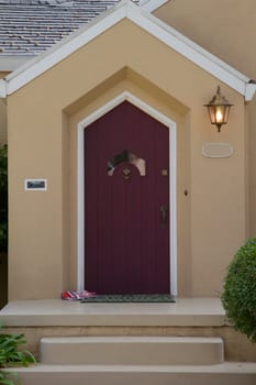 Burgandy door with unusual window on a white trimed beige hous with a lamp