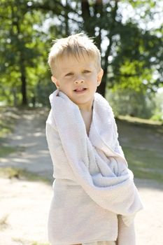 young blond boy wrapped in towel standing at river in forest. Summer time