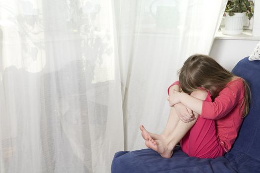 Little cute dreaming girl sitting on sofa at window hugging her knees. Child`s grief